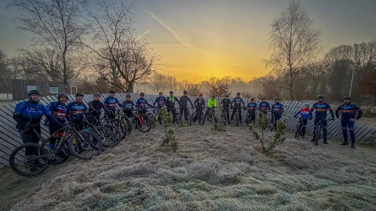 Groepsfoto van ruim twintig clubleden in clubkleding met hun mountainbikes op het bikepark, bij zonsopkomst