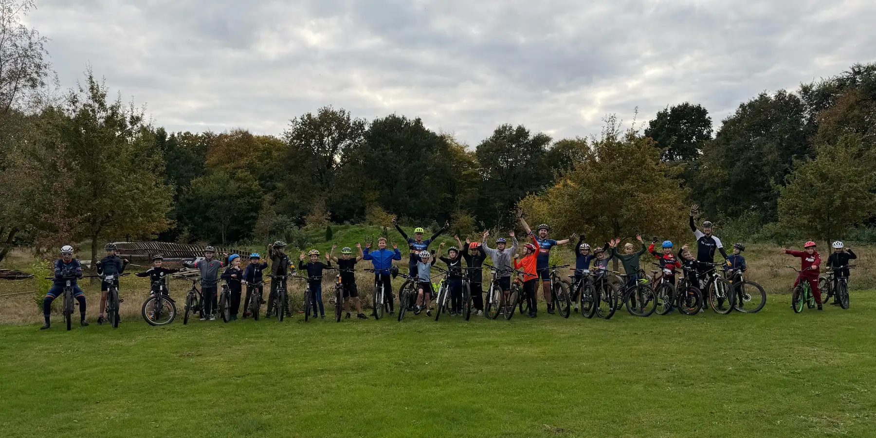 De jeugdgroep en hun trainers met hun mountainbikes op een grasveld, armen in de lucht